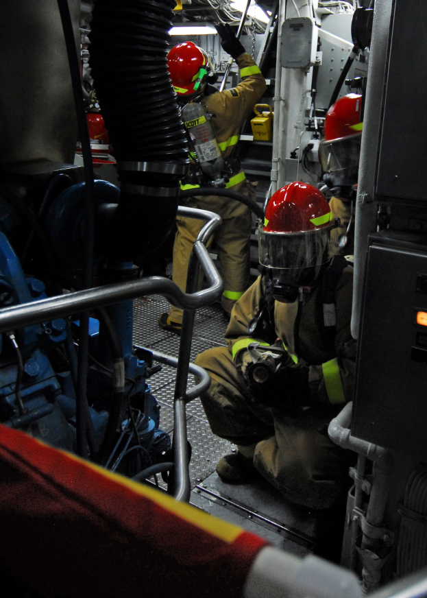 Feuerwehrleute in Helmen und Schutzausrüstung arbeiten an einem Feuerwehrauto mit Rohren und einer Lampe im Hintergrund.