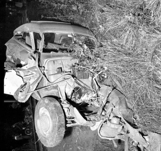 Ein Schwarz-Weiß- Foto eines schwer beschädigten Autos am Straßenrand, mit zerbeulter Front, zersplitterten Fenstern, einem Baum im Hintergrund und verstreuten Gegenständen um die Unfallstelle.