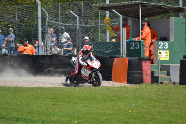 Eine Person in Helm und Handschuhen auf einem Motorrad auf einem grasbewachsenen Feld, mit Zuschauern, Absperrungen, Treppen und einem Schuppen im Hintergrund.