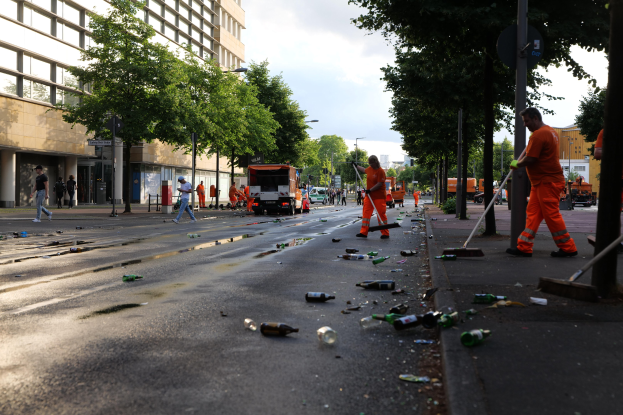 Menschen in orangen Uniformen sammeln Müll von einer Straße mit Flaschen und Schutt, mit Bäumen, Pfählen und Fahrzeugen im Hintergrund unter einem bewölkten Himmel.