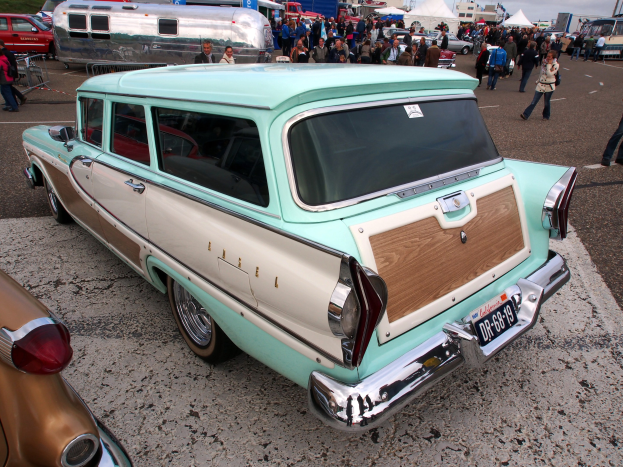 Ein grüner und weiße Stationwagen auf einem belebten Parkplatz mit Zelten, Masten, Bäumen und einem klaren blauen Himmel im Hintergrund.