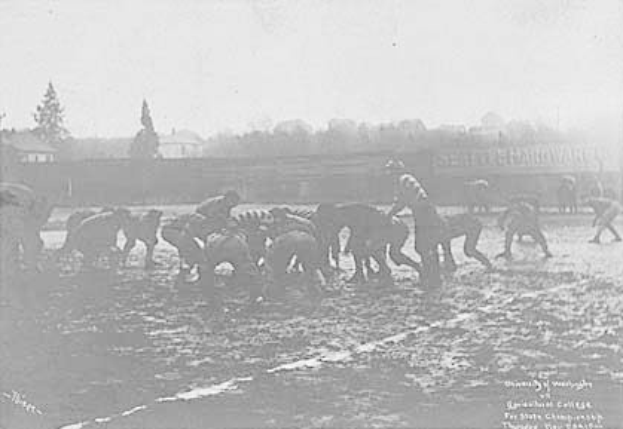 Ein Schwarz-Weiß-Foto einer Gruppe von Menschen, die auf einem Feld Fußball spielen, mit Pferden im Vordergrund und Bäumen, Gebäuden und Himmel im Hintergrund. Unten steht der Text "1918-1918 Fußball an der Staatsuniversität".