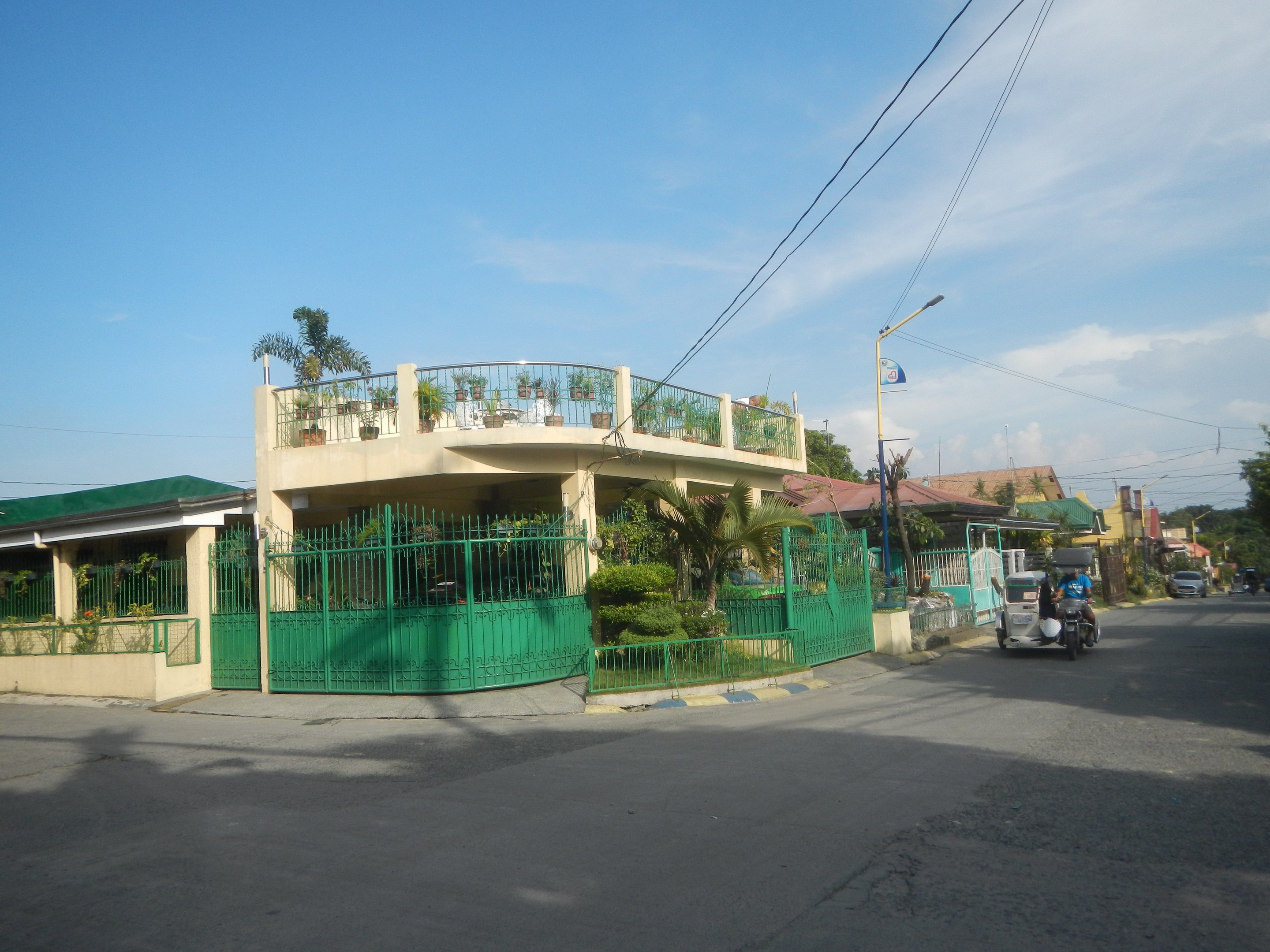 Eine Straße mit Fahrzeugen, ein grünes Gebäude mit einem Zaun, Geländern, Pflanzen, Bäumen, Polen, Lampen, Drähten, einer Flagge und einem bewölkten Himmel im Hintergrund.