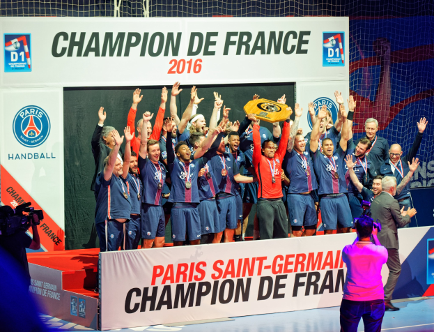 Eine Gruppe von Männern auf einer Bühne mit einem Pokal, einer Person, die filmt, und einem 'Champion de France 2016'-Schild im Hintergrund.