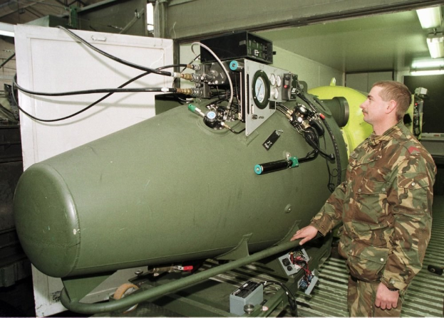 Ein Mann in militärischer Uniform steht neben einer großen grünen Bombe in einem unordentlichen industriellen Setting mit Maschinen, Kabeln und beleuchteten Lampen an der Wand hinter ihm, mit Text unten.