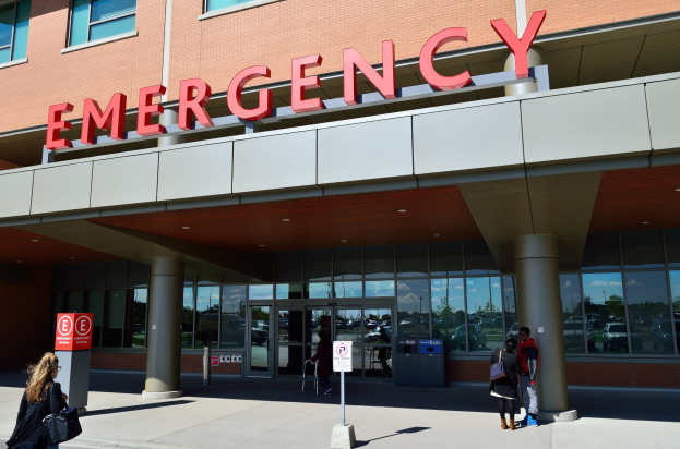 Eine Gruppe von Menschen geht vor einem Glaswand-Notfallgebäude mit Säulen, einem Namensschild, Hinweistafeln auf Pfählen und einem Mülleimer.