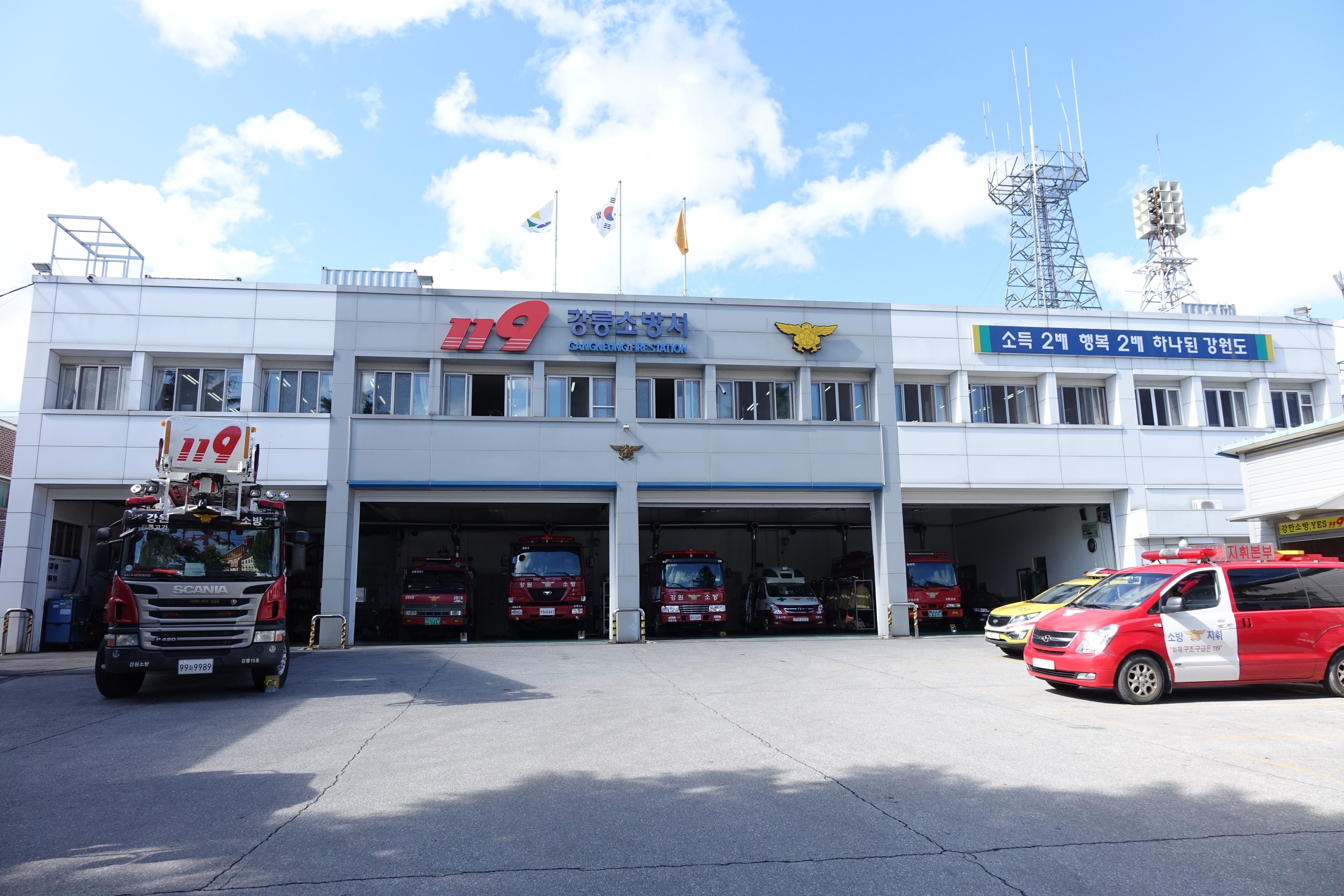 Feuerwache mit mehreren Feuerwehren vor dem Gebäude, das Fenster und Text aufweist, Fahnen an Stangen im Hintergrund, bewölkter Himmel und eine ferne Turm.