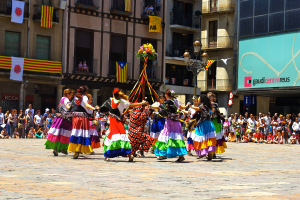 Eine Gruppe von Frauen in bunten Kleidern tanzt auf der Straße, umgeben von einer Menschenmenge, mit Gebäuden, Geländern, Balkonen, Fahnen, einem Laternenmast und einer Tafel mit Schrift im Hintergrund.