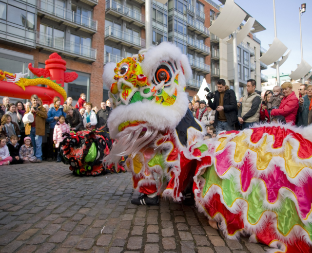 Ein lebendiges chinesisches Neujahrsfest in Amsterdam mit einer Löwen-Tanz-Performance vor einer Zuschauermenge, einige halten Kameras, vor dem Hintergrund von Gebäuden, Laternenmasten und einem klaren blauen Himmel.