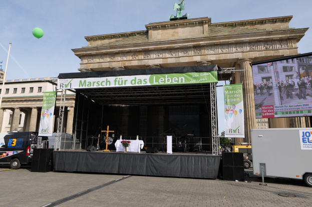 Bühne vor dem Brandenburger Tor mit einem Tisch, einer Vase, einem Kreuzsymbol, Fahnen, Lautsprechern, Fahrzeugen auf der Straße, Gebäuden mit Fenstern, einer Statue, einem Fahnenmast und einem bewölkten Himmel.