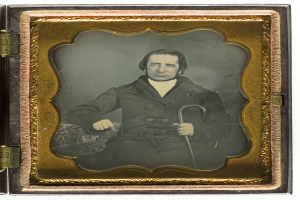Ein altes Daguerreotyp eines Mannes in einem Anzug, der in einem Stuhl sitzt und einen Stock hält, mit einem ernsten Gesichtsausdruck und konzentriertem Blick.