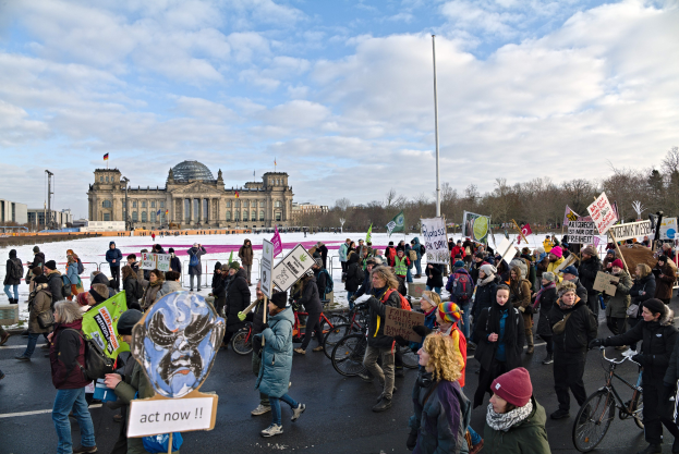 Eine große Gruppe von Menschen marschiert auf einer Berliner Straße und trägt Schilder und Banner, einige tragen Mützen oder Taschen, andere fahren Fahrräder, mit Wasser, Gebäuden, Bäumen, Polen und einem bewölkten Himmel im Hintergrund.