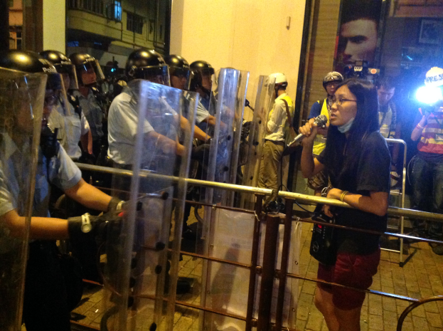 Eine Frau in schwarzem T-Shirt und roter Shorts steht vor einer Reihe von Bereitschaftspolizisten, die ein Mikrofon hält, umgeben von einer Gruppe von Menschen mit Helmen und Gegenständen in den Händen, mit einer Metallabsperrung dahinter und Gebäuden, Lichtern und einer Wand im Hintergrund.