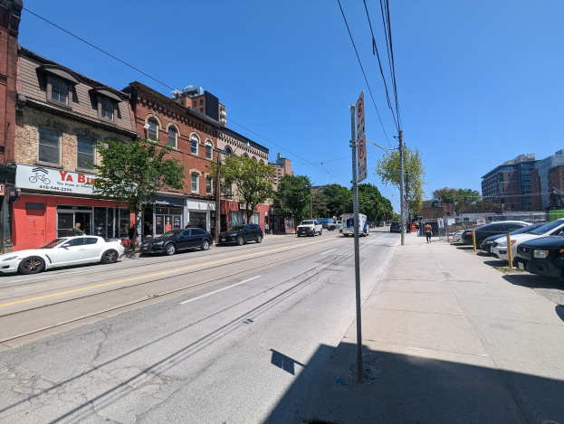 Stadtstraße mit parkenden Autos, Gebäuden, Bäumen, Strommasten mit Drähten und einem "New York City Real Estate for Sale"-Schild im Hintergrund vor einem sichtbaren Himmel.
