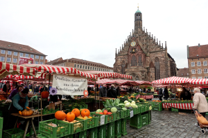 Ein belebter Markt in Nürnberg, Deutschland, mit verschiedenen Obst- und Gemüsesorten, Menschen, die umherlaufen, Zelten, Gebäuden mit Fenstern und einem Uhrturm im Hintergrund unter einem sichtbaren Himmel.