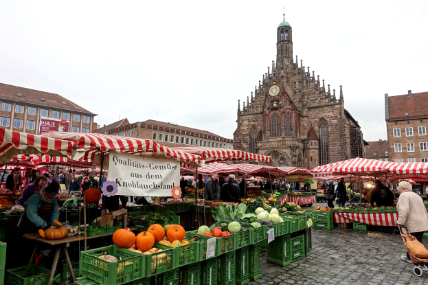Ein belebter Markt in Nürnberg, Deutschland, mit verschiedenen Obst- und Gemüsesorten, Menschen, die umherlaufen, Zelten, Gebäuden mit Fenstern und einem Uhrturm im Hintergrund unter einem sichtbaren Himmel.