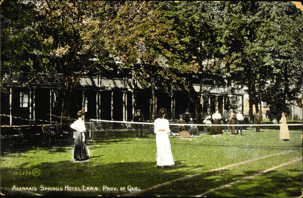 Eine Gruppe von Menschen, die Tennis auf einem Rasenplatz vor dem Abenakis Springs Hotel in Provo, Quebec, spielt, wobei einige Rackets halten, andere auf Stühlen sitzen, ein Netz in der Mitte und Bäume und ein Gebäude im Hintergrund, mit Text unten auf dem Bild.