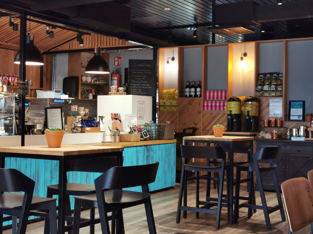 Innenraum eines Restaurants mit Holztischen, Stühlen, einer Topfpflanze, einer Schiefertafel, einem Kühlschrank, einem Feuerlöscher, Regalen mit Flaschen, Schränken mit Gegenständen, einem Rahmenfoto an der Wand, Deckenleuchten und Lampen.