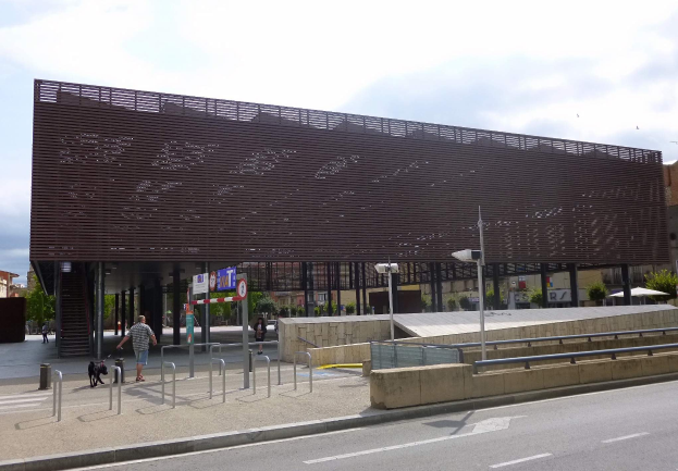 Großes modernes Gebäude mit zahlreichen Fenstern, das als neuer Eingang zur Stadtmitte dient und von Straßeninfrastruktur, Fußgängern, Fahrzeugen und Bäumen flankiert wird.