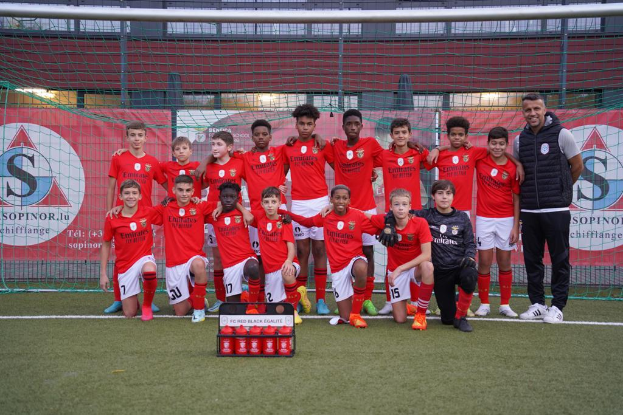 Eine Gruppe junger Männer in roten T-Shirts und schwarzen Shorts steht auf einem Fußballfeld neben einer Kiste roter Gegenstände, mit Schildern im Hintergrund, die 'FC Rot-Schwarz Eglaute - Champions League' lesen.
