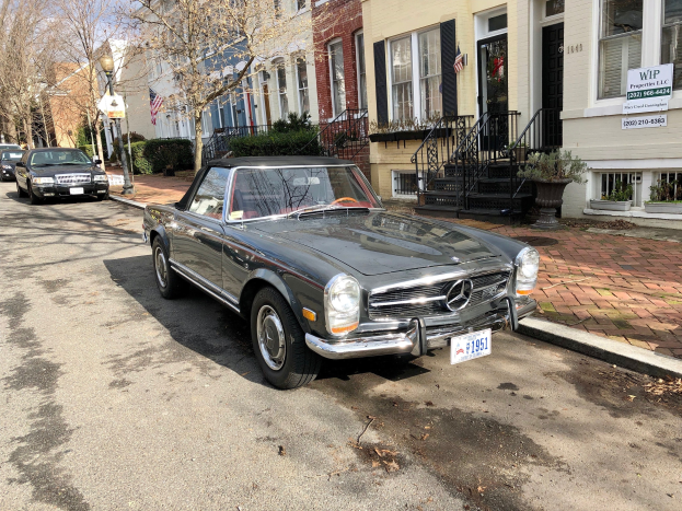 Ein 1971 Mercedes-Benz 280SL auf einer Stra├če mit Geb├Ąuden, Gel├Ąndern, T├Âpfen, B├Ąumen, Laternenmasten und Schildern im Hintergrund geparkt.