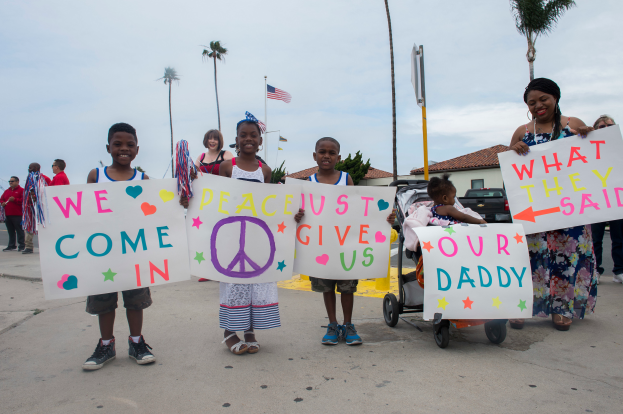 Eine Gruppe von Kindern hält ein Schild mit der Aufschrift "Wir kommen in Frieden, gebt uns unseren Daddy" und steht auf einer Straße umgeben von Bäumen, Polen, einer Fahne, einem Haus, Pflanzen und unter einem bewölkten Himmel, mit einer Frau, die einen Kinderwagen mit einem Baby darin schiebt.