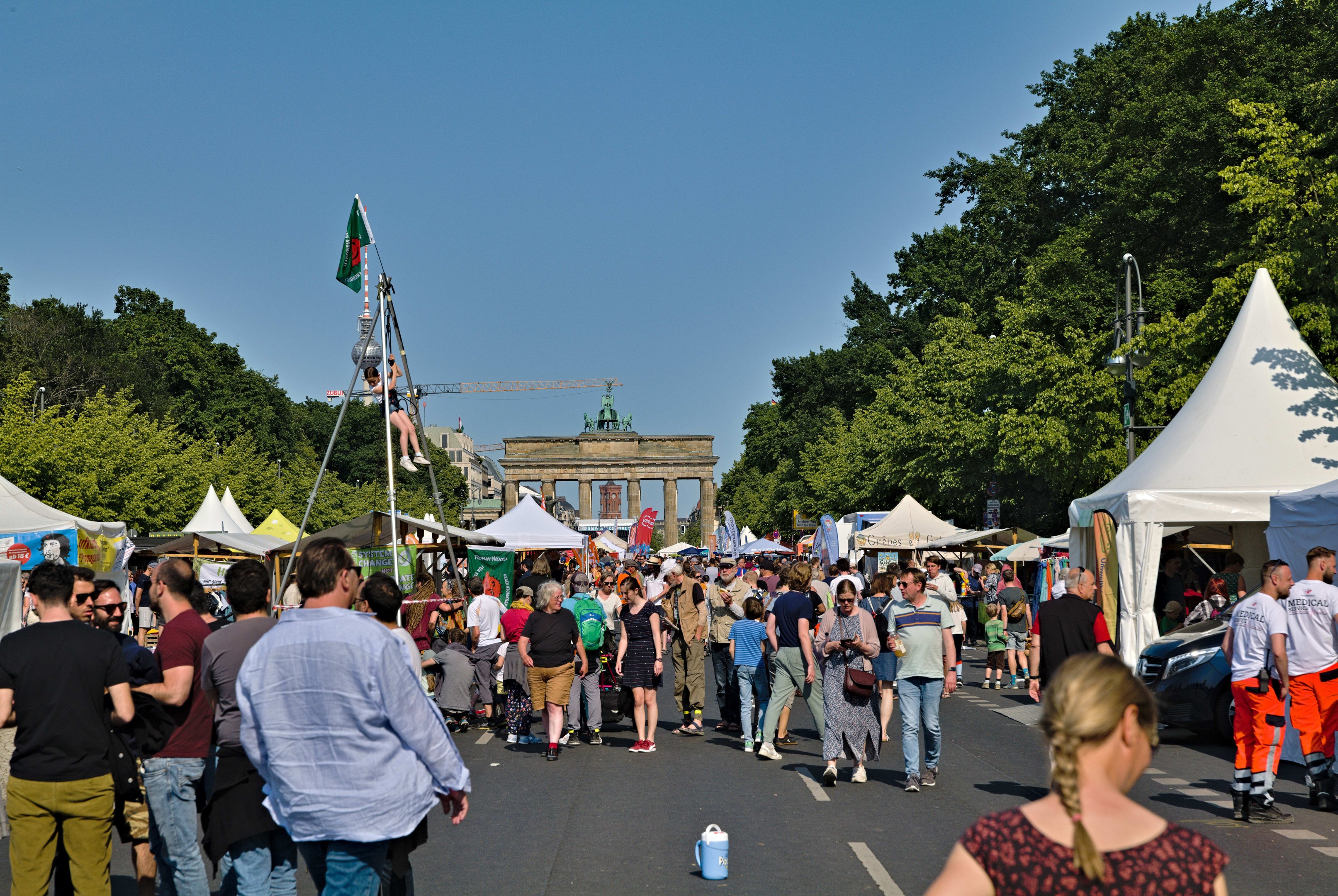 Eine Menschenmenge geht eine Straße mit Zelten, Fahrzeugen und Bäumen entlang, mit einem Bogen und einem klaren blauen Himmel im Hintergrund und Flaggenmasten auf der linken Seite, was auf das Oktoberfest in München, Deutschland, hindeutet.