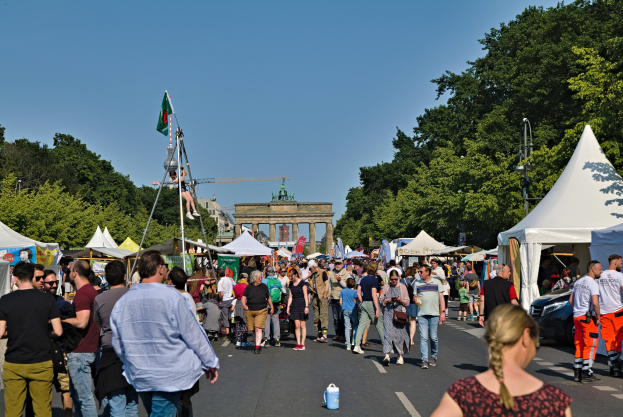 Eine Menschenmenge geht eine Straße mit Zelten, Fahrzeugen und Bäumen entlang, mit einem Bogen und einem klaren blauen Himmel im Hintergrund und Flaggenmasten auf der linken Seite, was auf das Oktoberfest in München, Deutschland, hindeutet.