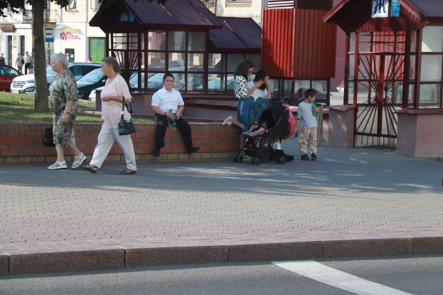 Eine Gruppe von Menschen, die mit einem Baby im Kinderwagen die Straße entlanggehen, umgeben von Gebäuden, Bäumen, Fahrzeugen und Gras, wobei einige Taschen tragen und ein Landfahrzeug im Hintergrund zu sehen ist.