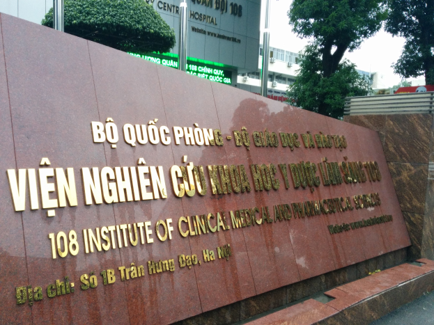 Übersicht des 108 Instituts für klinische Medizin und Pharmazie in Ho-Chi-Minh-Stadt, Vietnam, mit einer Textwand im Vordergrund, Bäumen, Versorgungsmästen, Gebäuden und einem klaren blauen Himmel im Hintergrund.