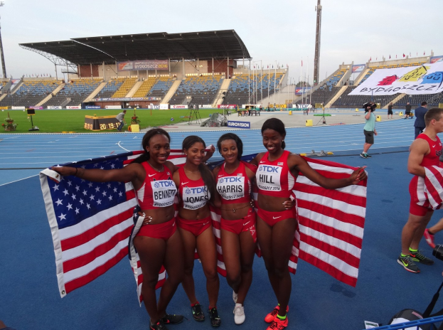 Vier Frauen in Sportkleidung stehen zusammen auf einer Laufbahn, lächeln und halten US-Flaggen, mit einem Stadion und Himmel im Hintergrund.