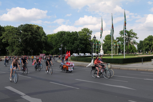 Eine Gruppe von Menschen, die auf Fahrrädern eine von Bäumen gesäumte Straße mit Laternenmasten und Flaggenmasten entlangfahren, während der Pariser Radtour 2016, mit bewölktem Himmel und ein paar Fahrzeugen im Hintergrund.