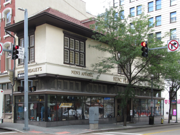 Eine belebte Stadtstraße mit einem geparkten Bus, Gebäuden, Bäumen, Laternenmasten, Verkehrszeichen und Schildern, mit einem Kaufhaus im Hintergrund unter einem sichtbaren Himmel.