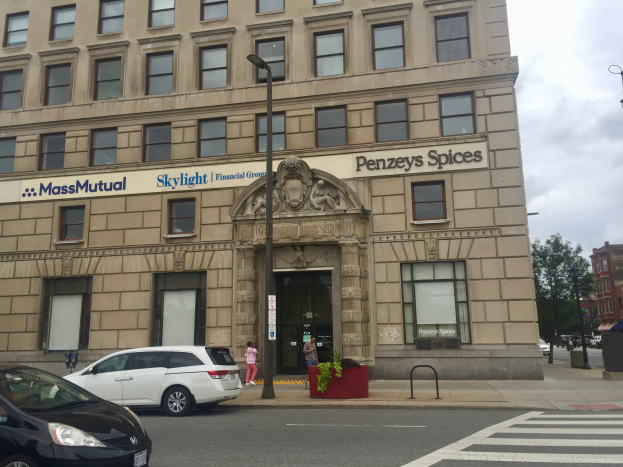 Großes Gebäude mit parkenden Autos, Straßenlaternen, Lichtern, fahrenden Fahrzeugen, Fußgängern auf dem Gehweg, Grünfläche und bewölktem Himmel, identifiziert als Hauptsitz der Massachusetts Mutual Financial Group.