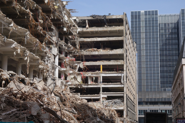 Ein große Gebäude wird in einer Stadt abgebrochen, umgeben von intakten Gebäuden, Straßeninfrastruktur, Fahrzeugen, Bäumen und einem klaren Himmel.