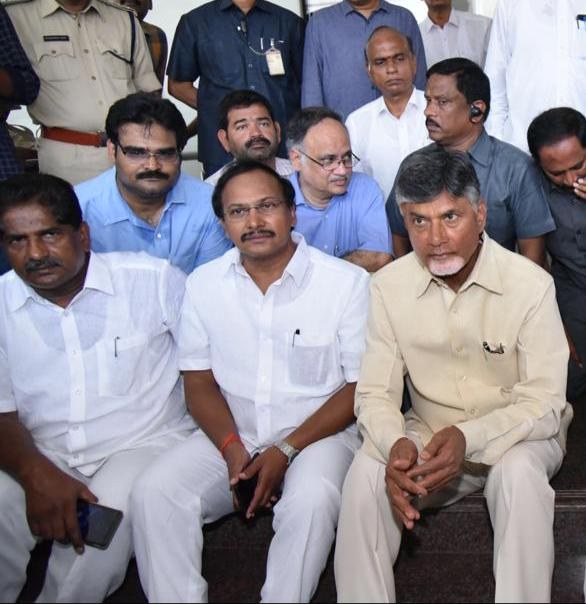 Chandrababu Naidu und sein Kabinett sitzen während einer Sitzung in einem öffentlichen Raum in Hyderabad, mit Menschen auf der Treppe und anderen im Hintergrund.