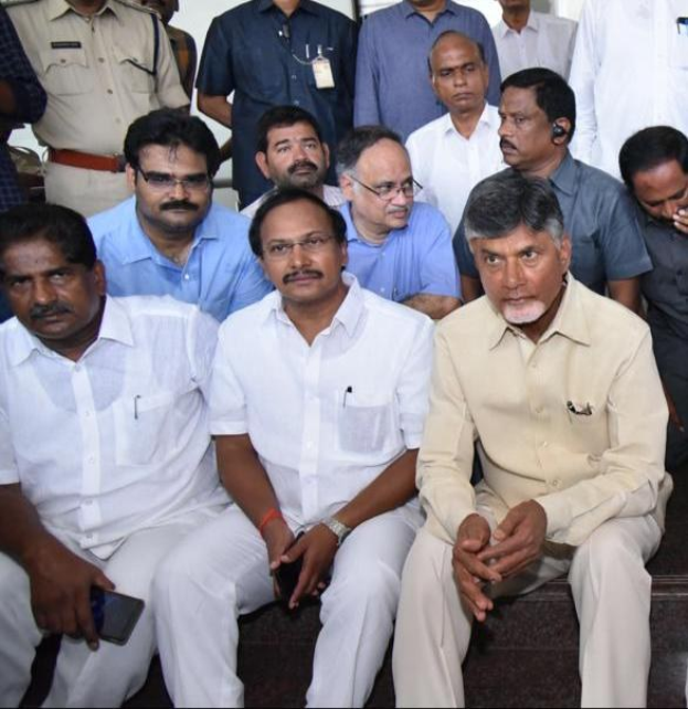 Chandrababu Naidu und sein Kabinett sitzen während einer Sitzung in einem öffentlichen Raum in Hyderabad, mit Menschen auf der Treppe und anderen im Hintergrund.