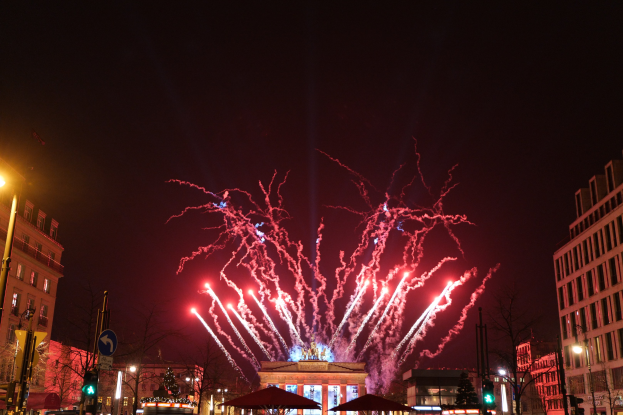 Eine belebte Stadtstraße in Berlin an Silvester, voller Menschen, Fahrzeuge und festlicher Dekoration, mit Feuerwerk, das den Nachthimmel erhellt.