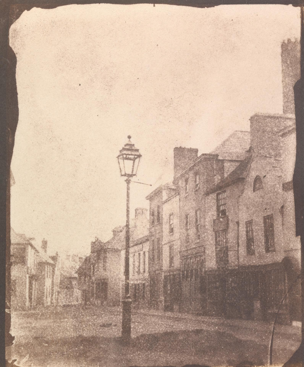 Altes Schwarz-Weiß-Foto einer zentralen Straßenlaterne zwischen Gebäuden unter einem klaren Himmel.