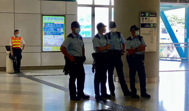 Eine Gruppe von Polizisten in Masken steht vor einem Gebäude mit Glastüren, Schildern mit Text, einer Säule und einem Geländer, wobei durch die Glastüren Bäume, Fahrzeuge und andere Gegenstände zu sehen sind.