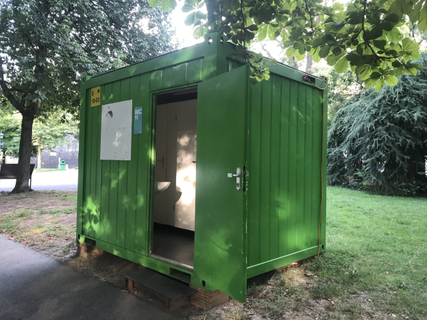 Grünes mobiles Toilettenhäuschen mit einer Tür und einer angebrachten Platte, das in einem Park mit Bäumen und einer Bank auf der linken Seite sowie dem Himmel im Hintergrund steht.