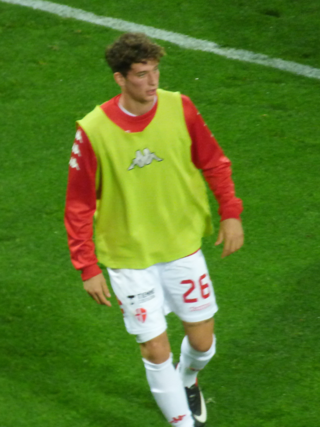 Ein Mann in einem gelb-roten T-Shirt läuft auf einem Fußballfeld, das mit weißen Linien markiert ist und trägt Fußballschuhe.