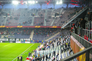 Ein großes Stadion voller Zuschauer, die ein Fußballspiel sehen, mit ein paar Personen in der Nähe des Feldes, einem Geländer und einer Fahne auf der rechten Seite und einem leeren Stadion mit Deckenbeleuchtung im Hintergrund.