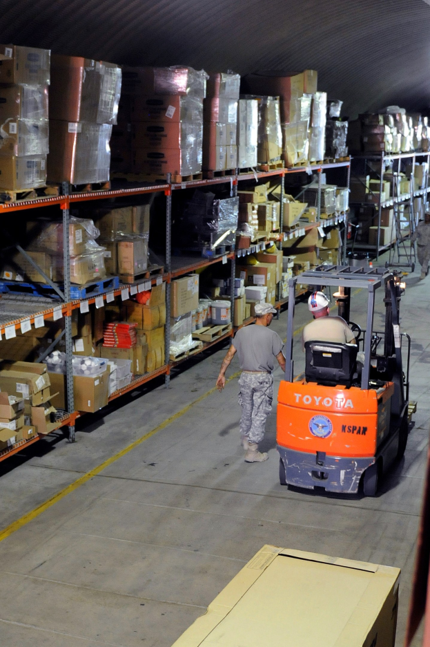 Ein Mann bedient einen Gabelstapler in einem Lagerhaus, das mit Kartons und Gegenständen auf Regalen gefüllt ist, mit einer Kiste am unteren Bildrand.