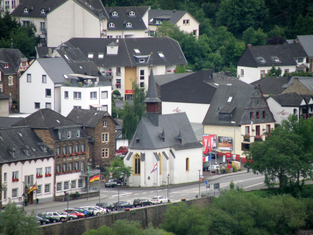 Eine kleine Stadt mit zahlreichen Gebäuden, Bäumen, Fahrzeugen und einer Kirche in der Mitte, mit Pflanzen und Wasser am unteren Bildrand.