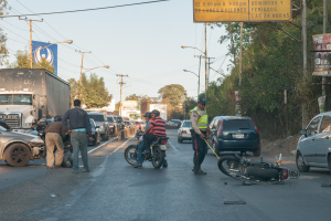 Eine Gruppe von Menschen um ein verunglücktes Motorrad auf der Straße herumstehend mit mehreren Fahrzeugen, einschließlich eines Lastwagens, und einem Hintergrund aus Bäumen, Pfählen, Lampen, Schildern und dem Himmel.