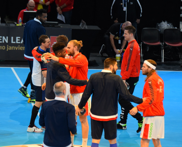Mannergruppe auf einem Futsal-Platz mit Bällen, die mit Stühlen, einem Tisch mit Flaschen und Laptops sowie einer Tafel mit Text im Hintergrund feiern.