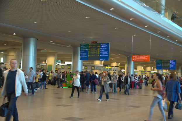 Menschen mit Gepäck durch den Flughafen gehen, überkopf-Schilder, Säulen, Deckenlampen und eine Glasbrüstung auf der rechten Seite.
