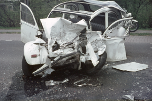 Ein Auto mit schwerer Frontschädigung steht am Straßenrand, mit Bäumen, einem Gebäude und einem weiteren Fahrzeug im Hintergrund.