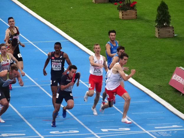 Eine Gruppe von Männern, die auf einer blauen Bahn laufen, umgeben von grünem Gras und Blumentöpfen, mit einer Texttafel auf der rechten Seite, die auf einen Wettbewerb hinweist, wahrscheinlich die Europameisterschaften im Leichtathletik.